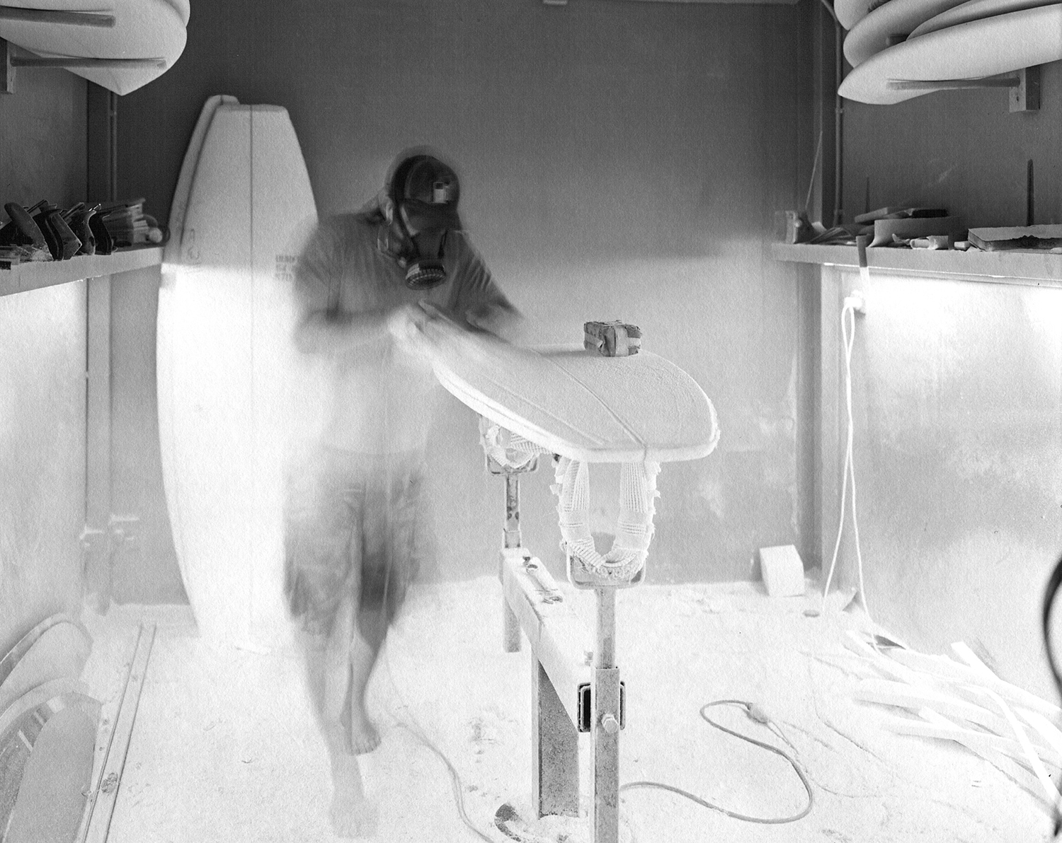 black and white image of man with mask shaping surfboard.